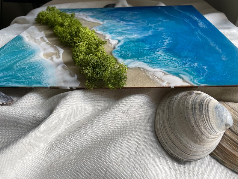 Pu&ograve; includere: Un'opera d'arte rettangolare raffigurante una scena di spiaggia con un oceano blu vibrante, onde bianche e schiumose e una striscia di sabbia. Il muschio verde rappresenta la terra. Una conchiglia si trova in primo piano, aggiungendo un elemento naturale al tema costiero.
