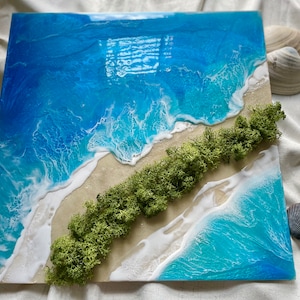 Pu&ograve; includere: Opera d'arte quadrata in resina raffigurante una scena di spiaggia. L'opera presenta vibranti onde blu e bianche che si infrangono su una riva sabbiosa, con una striscia diagonale di muschio verde. Conchiglie sono collocate attorno all'opera.