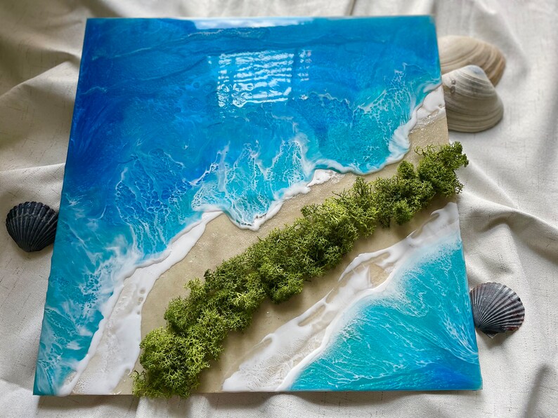 Pu&ograve; includere: Opera d'arte quadrata in resina raffigurante una scena di spiaggia. L'opera presenta vibranti onde blu e bianche che si infrangono su una riva sabbiosa, con una striscia diagonale di muschio verde. Conchiglie sono collocate attorno all'opera.