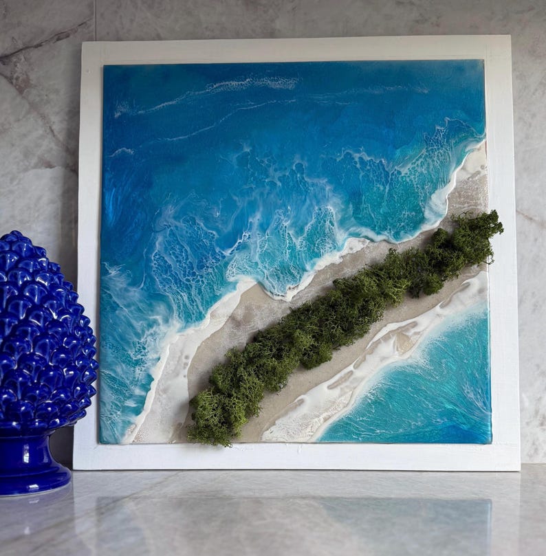 Pu&ograve; includere: Opera d'arte da parete quadrata raffigurante una scena oceanica in una cornice bianca. L'opera presenta un vivace oceano blu con onde bianche schiumose, una spiaggia sabbiosa e una striscia di muschio verde. Un pezzo unico di arredamento per la casa.