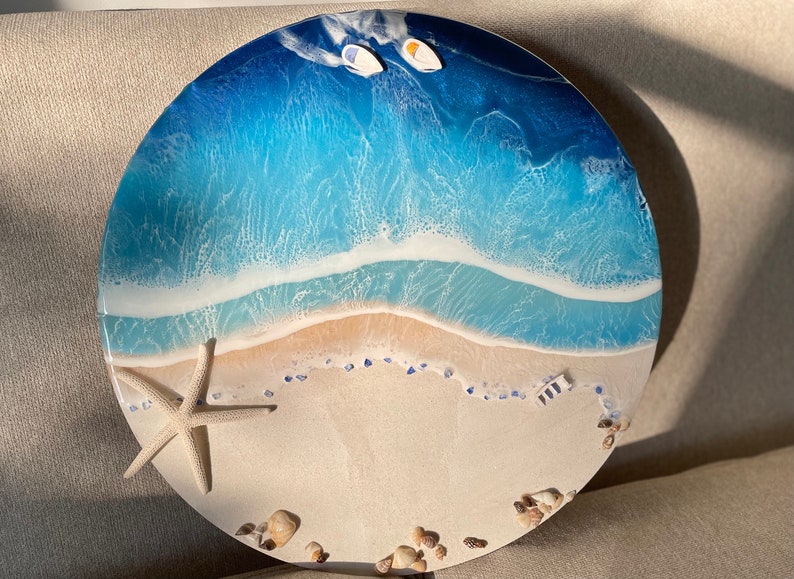 Pu&ograve; includere: Opera d'arte rotonda in resina raffigurante una scena di spiaggia. L'opera presenta una riva sabbiosa, onde schiumose in tonalit&agrave; di blu e due piccole barche. Una stella marina e conchiglie sono posizionate sulla sabbia. &Egrave; un oggetto decorativo.