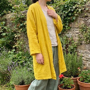 Könnte beinhalten: Ein langer, senfgelber Cardigan mit strukturiertem, geknittertem Aussehen. Der Cardigan wird über einem hellen Oberteil und mit grünen Hosen getragen. Der Hintergrund zeigt eine Steinmauer und Topfpflanzen, wodurch eine natürliche Umgebung entsteht.