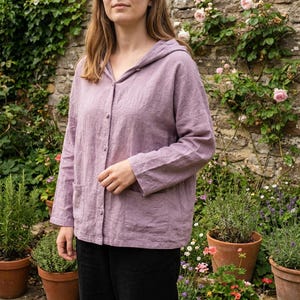Könnte beinhalten: Eine hellviolette Kapuzenjacke aus Leinen mit langen Ärmeln, einer Knopfleiste und zwei Vordertaschen. Die Jacke wird mit einer schwarzen Hose getragen. Der Hintergrund zeigt eine Steinmauer und Topfpflanzen.