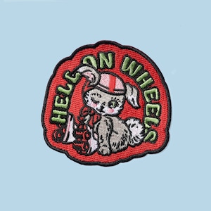 以下が含まれることがあります： 赤い刺繍のパッチ。ヘルメットをかぶって赤と白の縞模様のものを抱えた漫画のウサギが描かれています。ウサギの周りに緑色の文字で「HELL ON WHEELS」と書かれています。