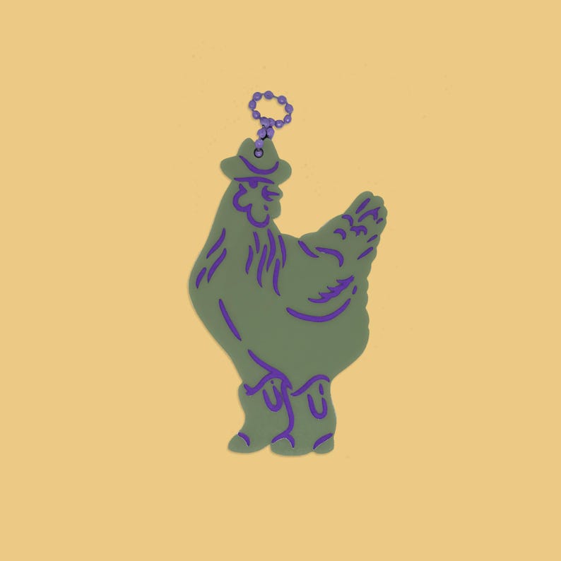 May include: A green chicken-shaped keychain with purple accents, wearing a cowboy hat and boots. The keychain has a purple ring for attachment. The background is a solid yellow color.