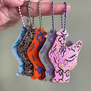 May include: A collection of colorful rooster-shaped keychains. Each keychain features a unique design, including glitter and various color schemes such as blue, brown, orange, gray, and pink. They are attached to ball chains.