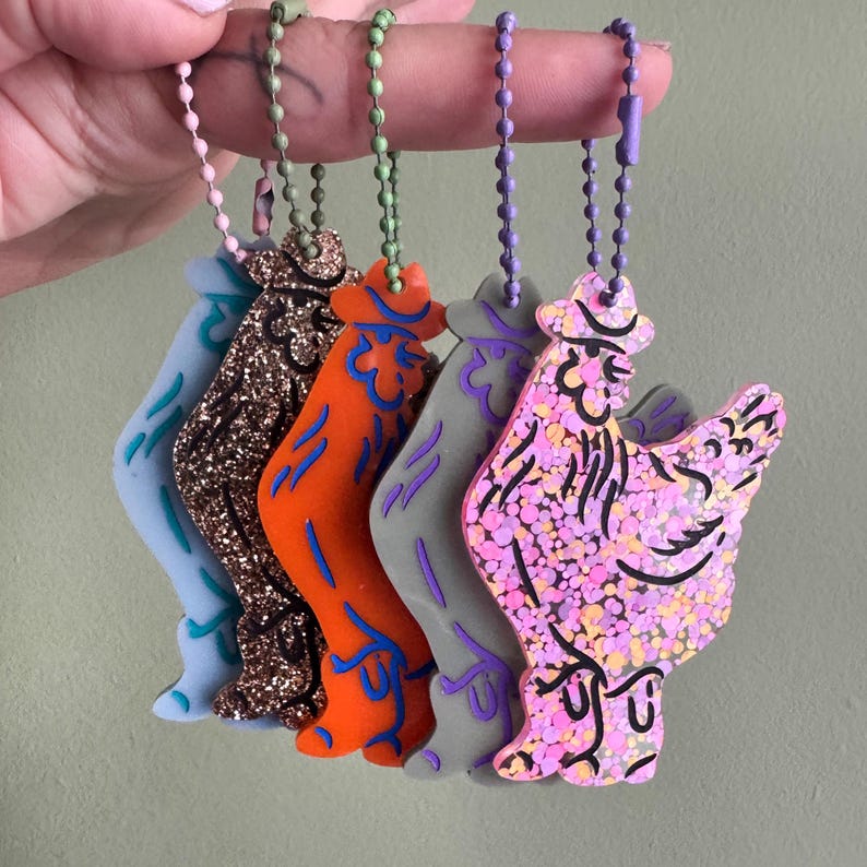 May include: A collection of colorful rooster-shaped keychains. Each keychain features a unique design, including glitter and various color schemes such as blue, brown, orange, gray, and pink. They are attached to ball chains.