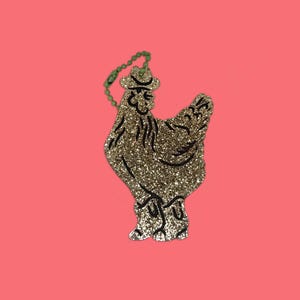May include: Glittery gold chicken-shaped ornament with black details, wearing a hat, and a green beaded chain. The ornament is set against a solid pink background. A fun, decorative item.