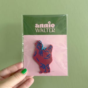 May include: A maroon and blue chicken-shaped pin on a pink card, packaged in a clear plastic sleeve. The card has the text "annie WALTER" in white letters on a green background. The pin is held by a hand with a green painted nail.