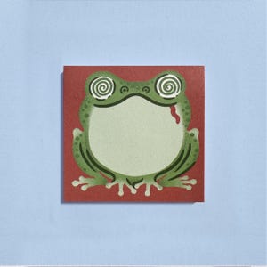May include: A green frog illustration with spiral eyes and a red mouth, set against a red square background. The frog is outlined in dark green, with a light green body and a white belly. The background is on a light blue surface.