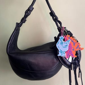 May include: Black leather crescent-shaped handbag with a braided strap. The bag is adorned with colorful, cartoonish keychains in blue, pink, and orange. The bag hangs against a light green background.
