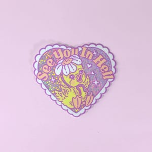 Op de afbeelding: Een hartvormige sticker met een geschulpte rand, met een gele kuiken met een madeliefje en de tekst "See You In Hell". De sticker heeft een glitterachtig, holografisch effect met roze en paarse tinten.