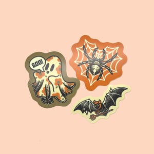 May include: Three Halloween stickers: a ghost with a floral pattern and a speech bubble saying "BOO!", a spider on a web, and a bat with a bow. The stickers have a vintage style.