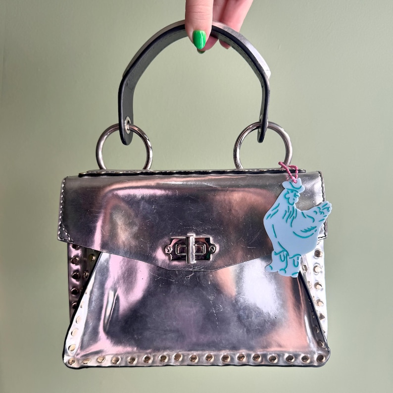 May include: A silver metallic handbag with a top handle and metal ring attachments. The bag has a rectangular shape with a clasp closure and is adorned with rivets. A light blue chicken-shaped charm hangs from the handle.