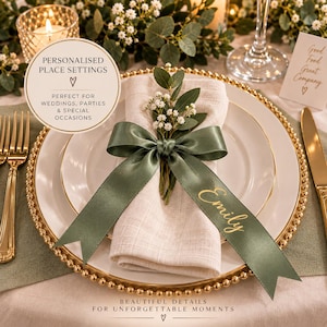 May include: A white dinner plate with a gold rim features a folded beige linen napkin tied with a sage green satin ribbon. The ribbon is printed with the name Emily in gold script and decorated with a small sprig of white baby's breath flowers.