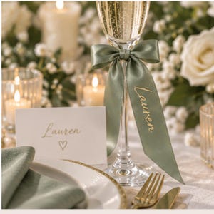 May include: A clear glass champagne flute adorned with a sage green satin ribbon tied in a bow, featuring the name Lauren printed in gold script. A matching white place card with the name Lauren and a small heart icon in gold script is included.