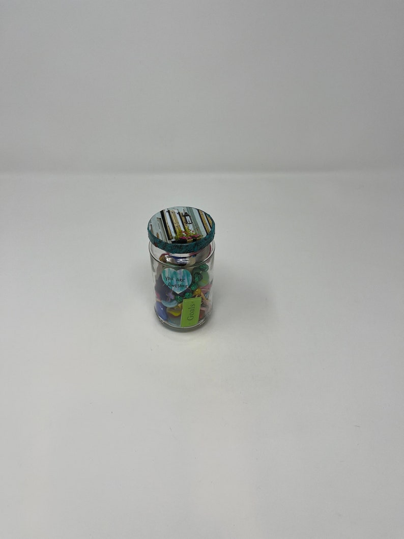 Pode incluir: Um frasco de vidro com uma tampa listrada azul-esverdeada e branca cheio de bugigangas coloridas e pequenos bilhetes de papel. Os bilhetes t&ecirc;m mensagens como "Voc&ecirc; &eacute; incr&iacute;vel" e "Metas".