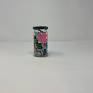 Pode incluir: Um frasco de vidro transparente com tampa azul-esverdeada cheio de contas coloridas, bot&otilde;es e um cora&ccedil;&atilde;o de feltro rosa.