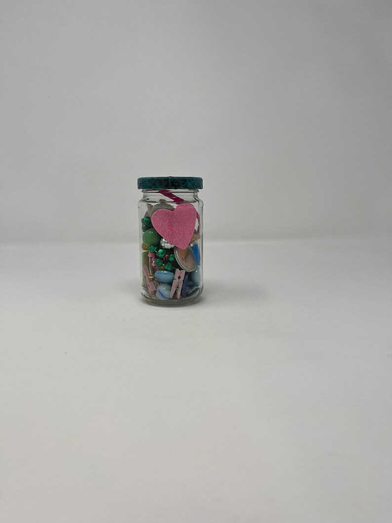 Pode incluir: Um frasco de vidro transparente com tampa azul-esverdeada cheio de contas coloridas, bot&otilde;es e um cora&ccedil;&atilde;o de feltro rosa.