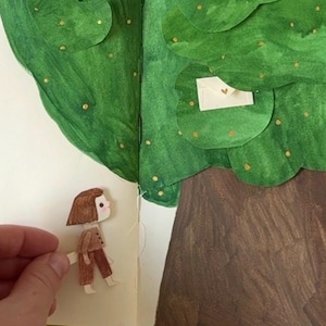 Op de afbeelding: Een handgemaakte papierkunstscène met een boom met een bruine stam en groene bladeren met gouden accenten. Een kleine papieren figuur is bij de boom geplaatst. Een rode roos staat op de achtergrond.