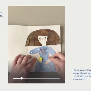 May include: A handmade paper doll with a blue dress and brown hair is displayed in an open book. The doll holds a yellow star. Text on the left reads "Now, let's open your story and see how it came to life."