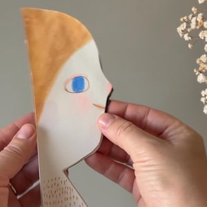 Può includere: Ritaglio di carta di un profilo con capelli arancioni, un occhio blu e una guancia rosa. Il ritaglio è tenuto tra due mani. Lo sfondo è grigio chiaro, con fiori bianchi nell'angolo in alto a destra.