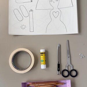 May include: A craft supplies arrangement featuring a paper doll outline, a roll of tape, glue stick, craft knife, scissors, a box of colored pencils, and small fasteners. The paper doll has a heart-shaped detail.