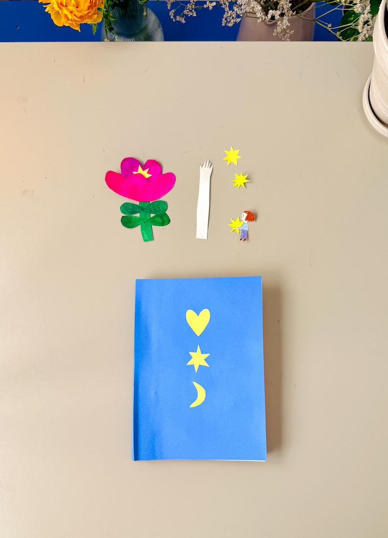 May include: A blue notebook with yellow heart, star, and crescent moon cutouts. Paper cutouts of a pink flower, white arm, yellow stars, and a person holding a star are scattered around the notebook. The image is taken from above.