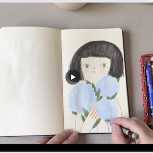 May include: An open sketchbook with a drawing of a girl holding blue flowers. A hand is coloring the drawing with a green crayon. Colored pencils and crayons are in trays on either side of the sketchbook.