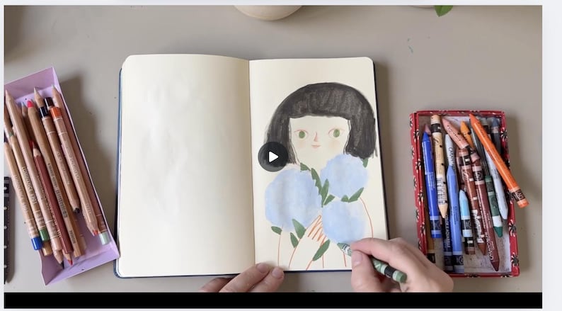 May include: An open sketchbook with a drawing of a girl holding blue flowers. A hand is coloring the drawing with a green crayon. Colored pencils and crayons are in trays on either side of the sketchbook.