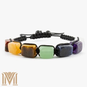 May include: A chakra bracelet featuring seven colorful, rectangular gemstones. The stones are strung on a black, braided cord with an adjustable sliding knot closure. The gemstones include red, yellow, brown, green, blue, purple, and white.