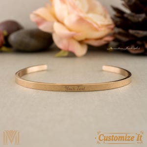 May include: A rose gold cuff bracelet with the engraved text "Your Text". The bracelet is a simple, open design. The background includes a blurred peach-colored flower and a pine cone.