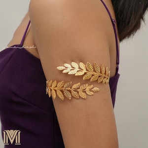 May include: Gold-colored arm cuff jewelry in a leaf design. The jewelry is worn on the upper arm and is made of a shiny, metallic material. The leaves are detailed and arranged in a symmetrical pattern. The background is a solid purple color.