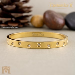 May include: A gold-coloured bangle bracelet featuring star-shaped designs, each set with a small, clear gemstone. The bracelet is engraved with the text "Your Text". The words "Customize It" are also visible.