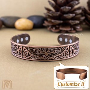 May include: A copper-toned cuff bracelet featuring an embossed tree of life design and intricate Celtic knotwork patterns. The interior of the bracelet displays small circular magnets. An inset shows the smooth inner band with the text Customize It.