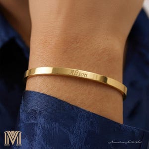 Può includere: Un bracciale a polsino dorato inciso. Il bracciale è una fascia semplice e lucida con il nome "Alison" inciso sul davanti. Il bracciale è mostrato sul polso di una persona, su uno sfondo blu scuro.