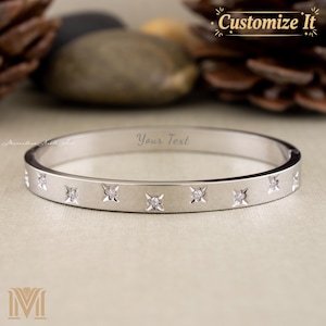 May include: A silver-colored bangle bracelet with a series of small, sparkling diamond-like accents. The bracelet has the text "Your Text" engraved on it. The words "Customize It" are also visible.