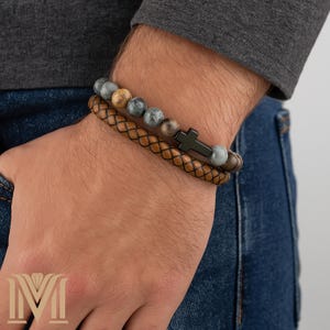 May include: Three bracelets are visible on a wrist. The top bracelet has grey and brown stone beads. The middle bracelet is woven brown leather. The bottom bracelet has a black cross charm and grey stone beads. The person is wearing a grey long-sleeve shirt and blue jeans.