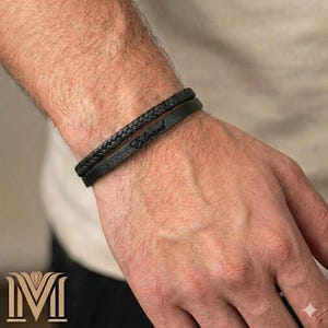 Peut inclure: Deux bracelets en cuir noir. L'un est tressé, l'autre est une bande simple avec le nom "Edward" gravé. Les bracelets sont portés au poignet, mettant en valeur un accessoire classique et personnalisé.