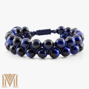 May include: A double-strand bracelet featuring alternating black and blue tiger eye beads. The beads are strung on a dark blue cord, creating a woven pattern. The bracelet has an adjustable sliding knot closure.