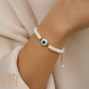 May include: White beaded bracelet with a gold evil eye charm. The charm has a blue eye with a white pupil and a gold rim.