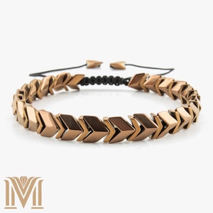 Könnte beinhalten: Ein bronzefarbenes Armband mit Pfeilform. Das Armband ist an einem schwarzen Kordelband mit verstellbaren Enden befestigt. Die pfeilförmigen Perlen sind metallisch und in einem sich wiederholenden Muster angeordnet.