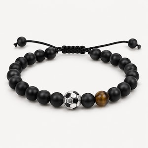 May include: A beaded bracelet featuring matte black round beads, a single silver-toned soccer ball bead, and one brown tiger eye bead. The bracelet is constructed with a black cord and an adjustable sliding knot closure with two small black beads.