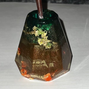 Puede incluir: Un portaplumas de resina verde y marrón con motas doradas. El portaplumas tiene forma de cristal geométrico y tiene una abertura redonda en la parte superior.