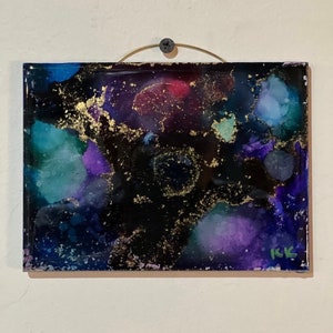 May include: Abstract art painting with a black background and swirling colours of purple, blue, green, and gold. The painting is framed with a thin gold frame and has the artist's initials "IK" in the bottom right corner.