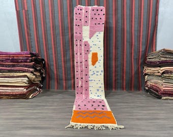May include: A long, rectangular rug with a geometric pattern in shades of pink, orange, white, and black. The rug has a fringe border and is made of wool.
