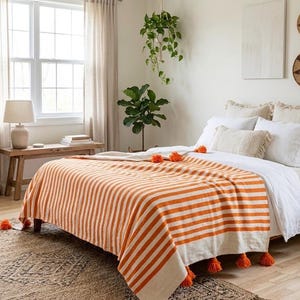 May include: An orange and white striped throw blanket with orange tassels on a bed. The blanket is draped over a bed with white bedding. The room has a neutral color palette with a wooden bench and a lamp.