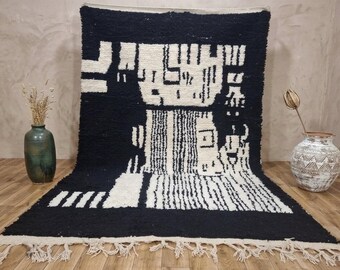 May include: A rectangular area rug featuring a high-pile texture with a bold black and white geometric pattern. The design consists of abstract shapes and lines, finished with a fringe along the bottom edge. The rug is displayed in an upright position.