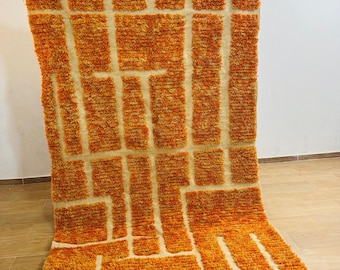 May include: A rectangular, orange and beige rug with a geometric pattern. The rug features a shaggy texture and a design of orange rectangles and lines against a beige background. The rug has a fringed edge.