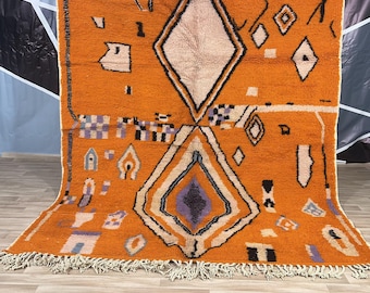 May include: A large, orange Moroccan rug with a geometric pattern in shades of black, white, and purple. The rug has a fringe border.